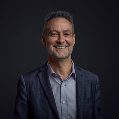 Glenn King wearing a jacket and collared shirt smiling broadly in front of a black background