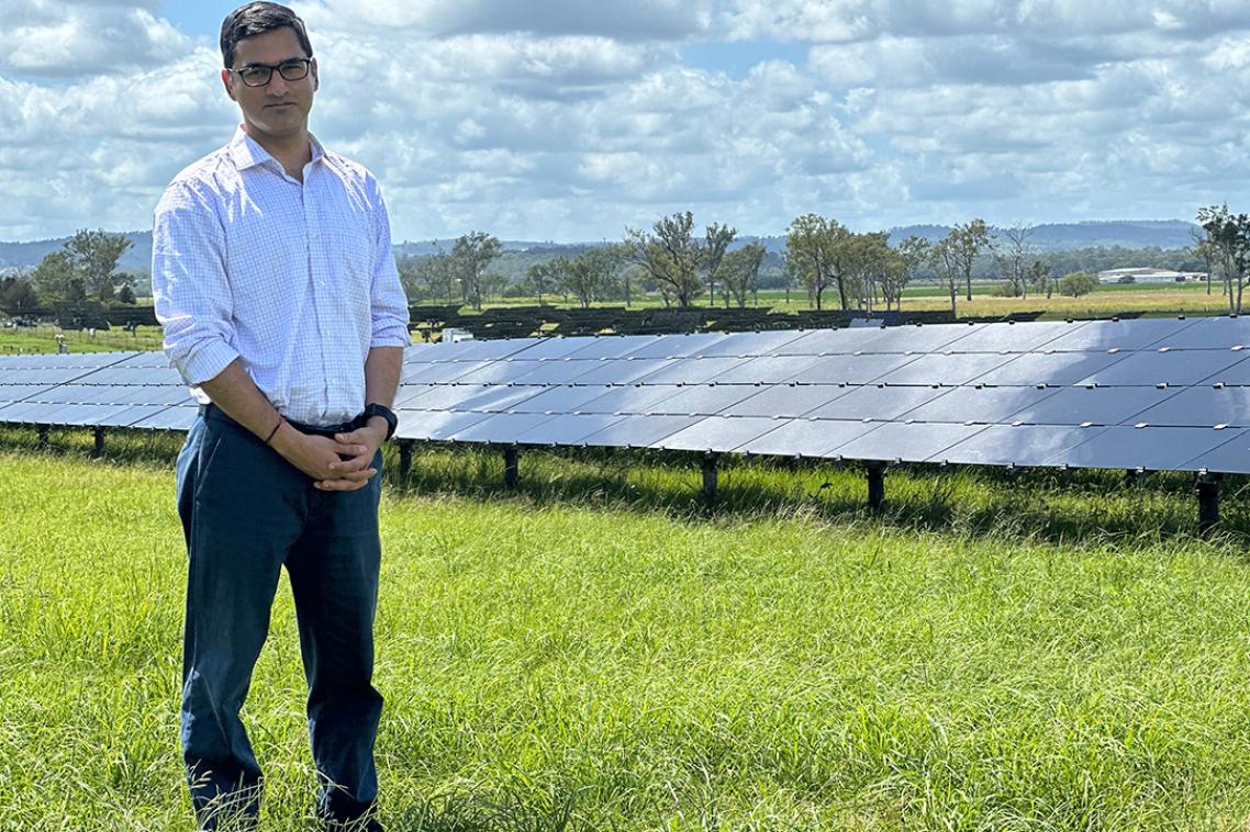 A man stands at a solar farm with dozens of solar panels behind him.